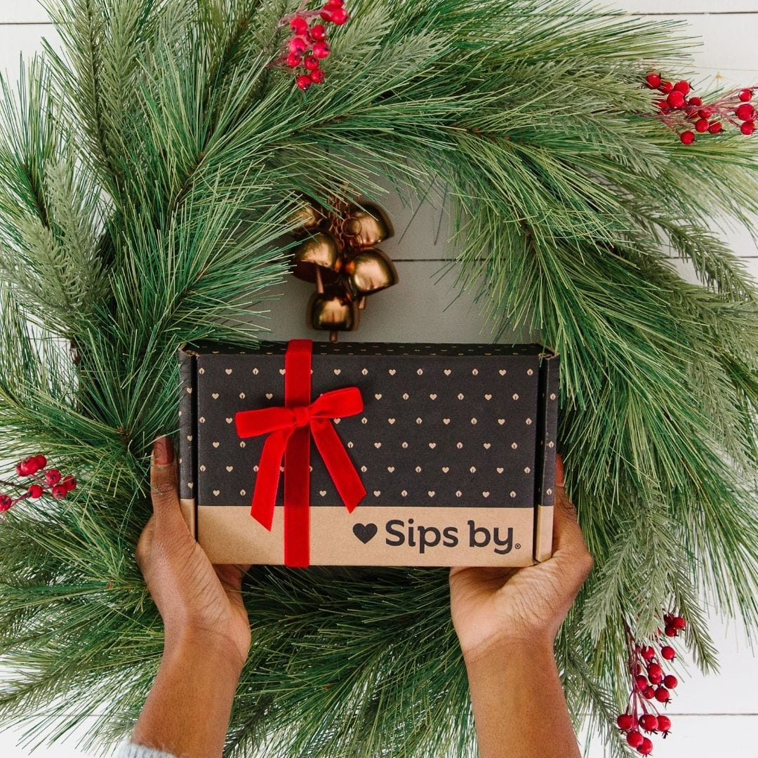 Holiday wreath with hands holding up a Sips by box wrapped in a red bow