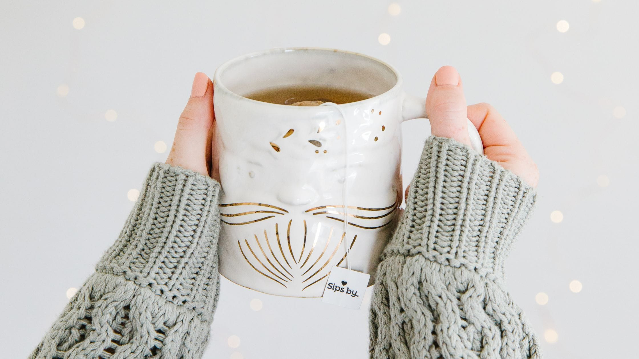 Green sweater wrapped sleeves holding up a white and gold Santa mug with Sips by tea
