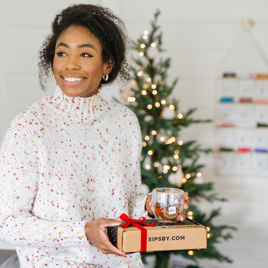 Smiling woman holding a Sips by Box and glass mug with a Christmas tree and Sips by Holiday Tea Advent Calendar in the background