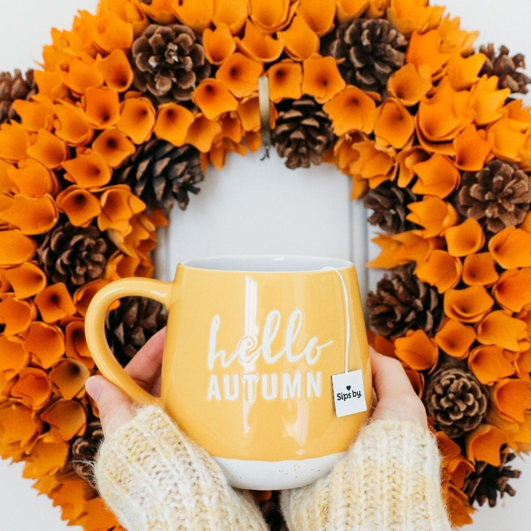 Autumn orange pinecone wreath with hands holding up a yellow hello autumn mug with Sips by tea