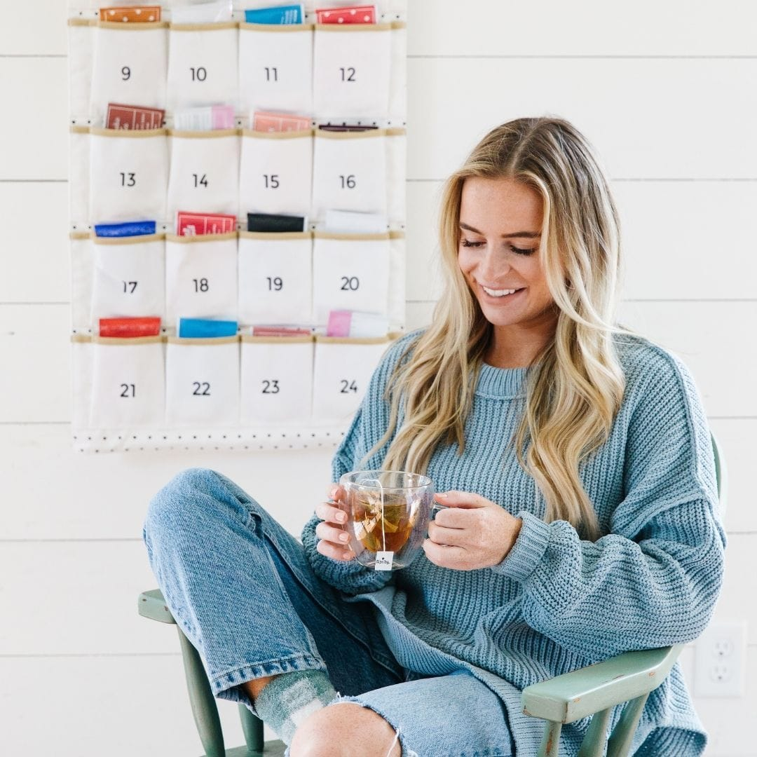 Woman sitting in a chair with the Sips by 24-day advent tea calendar behind her on the wall and holding a cup of advent tea
