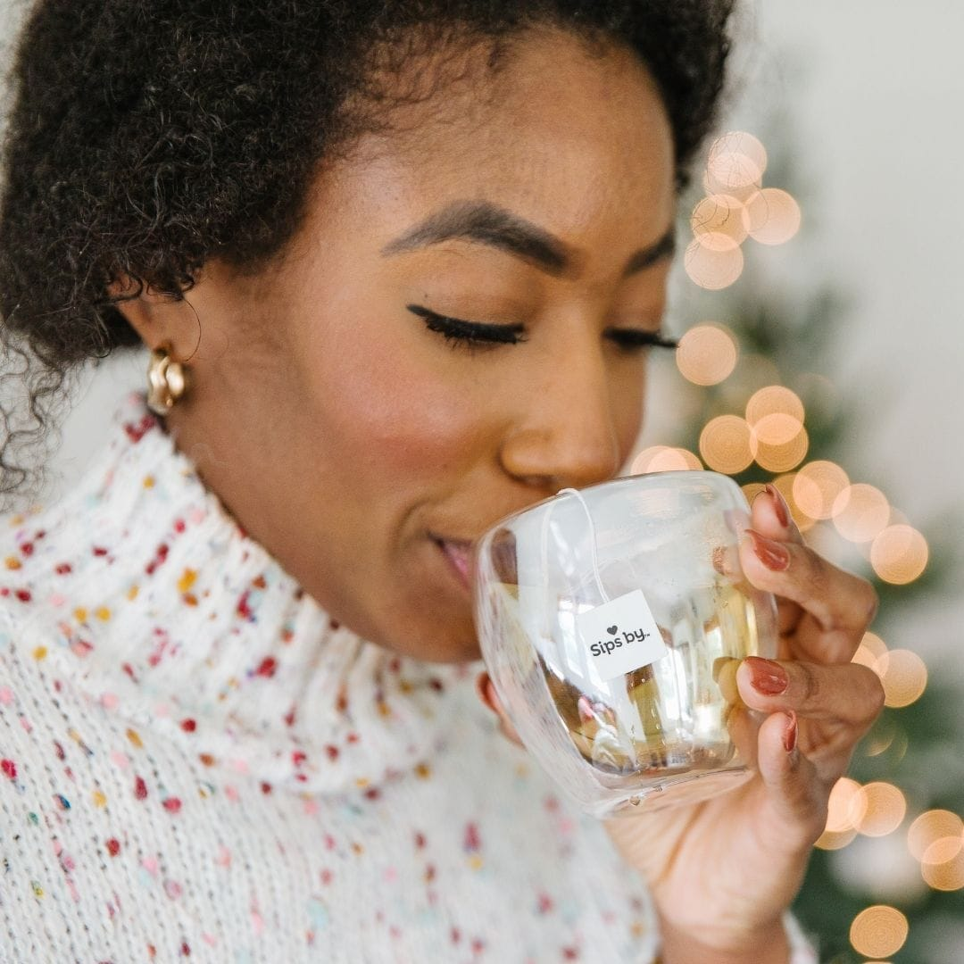 Woman drinking from a glass mug of Sips by tea with bokeh lights in the background