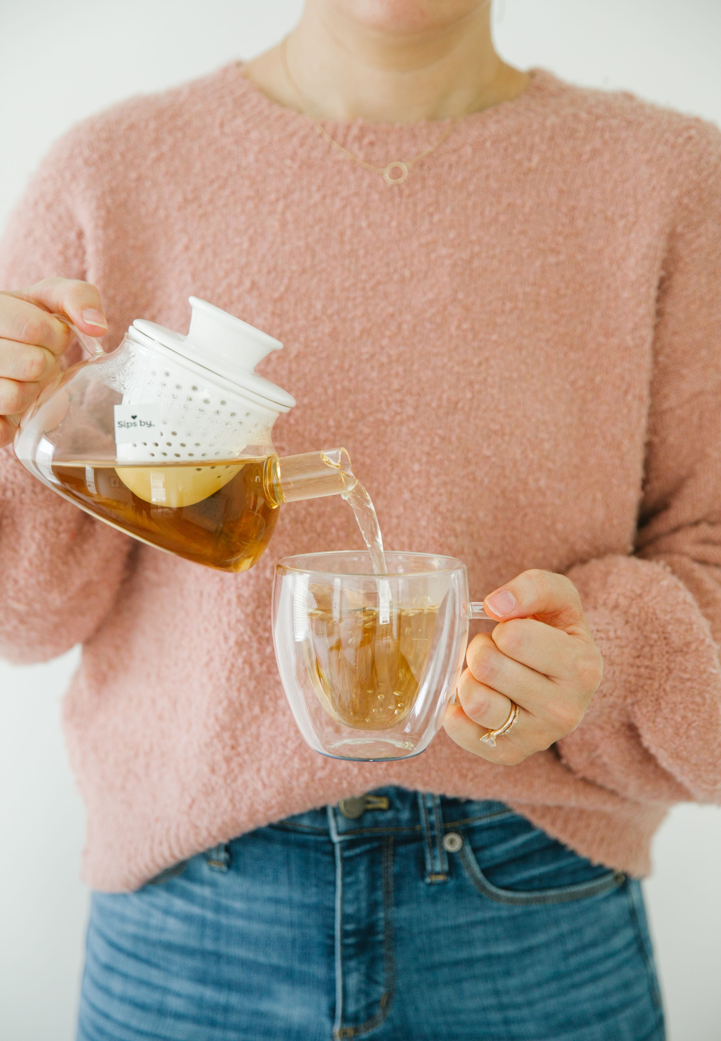 Person in a pink sweater pouring a glass teapot with an infuser filled with tea into a double-wall glass heart mug