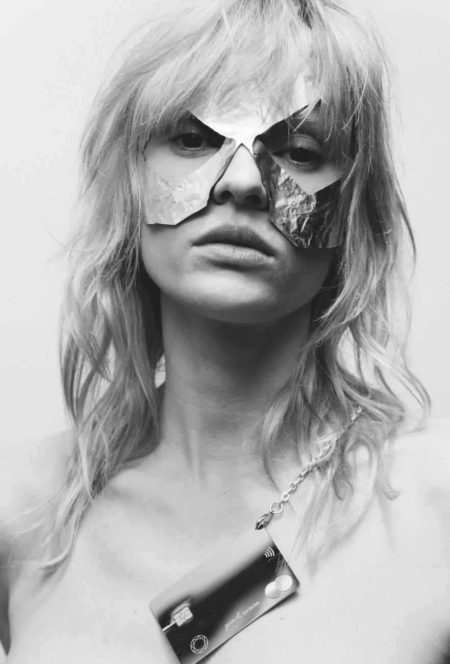 Black and white image for a masked girl wearing Saskia Diez Lovers card on her neck. 