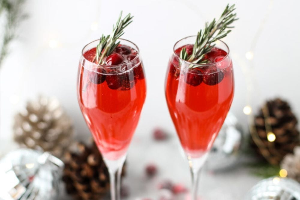 Winter Mimosas recipe with two glasses of red mimosas with cranberries, rosemary, and pinecones in the background - Serve these seasonal mimosas at your next holiday party for a festive pop of color. Merry mimosas, everyone! Read more.