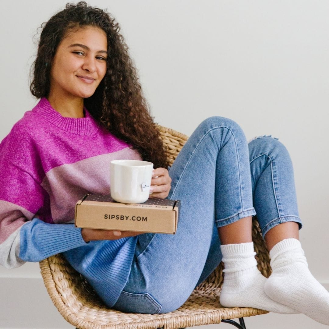 Picture of a black woman in a purple and blue striped sweater sitting in a chair and holding a Sips by Box with a mug resting on top