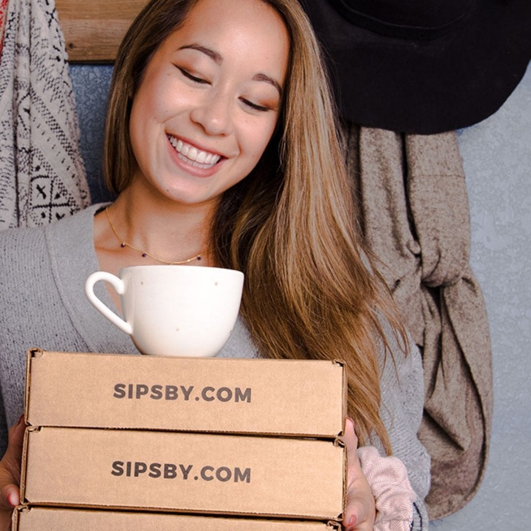Smiling woman holding a stack of Sips by Boxes with a white tea cup balanced on top
