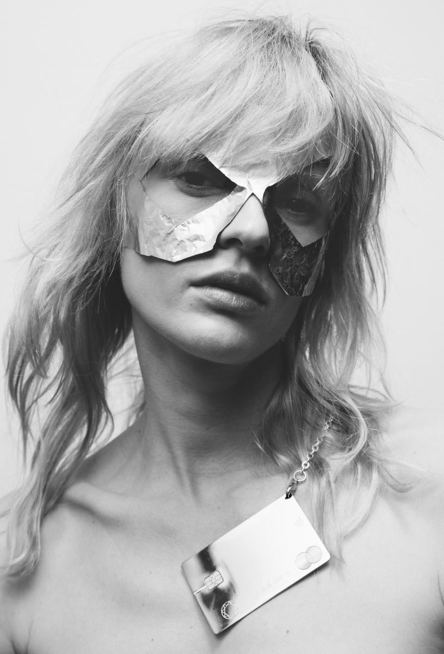 Black and white image for a masked girl wearing Saskia Diez Lovers card on her neck. 