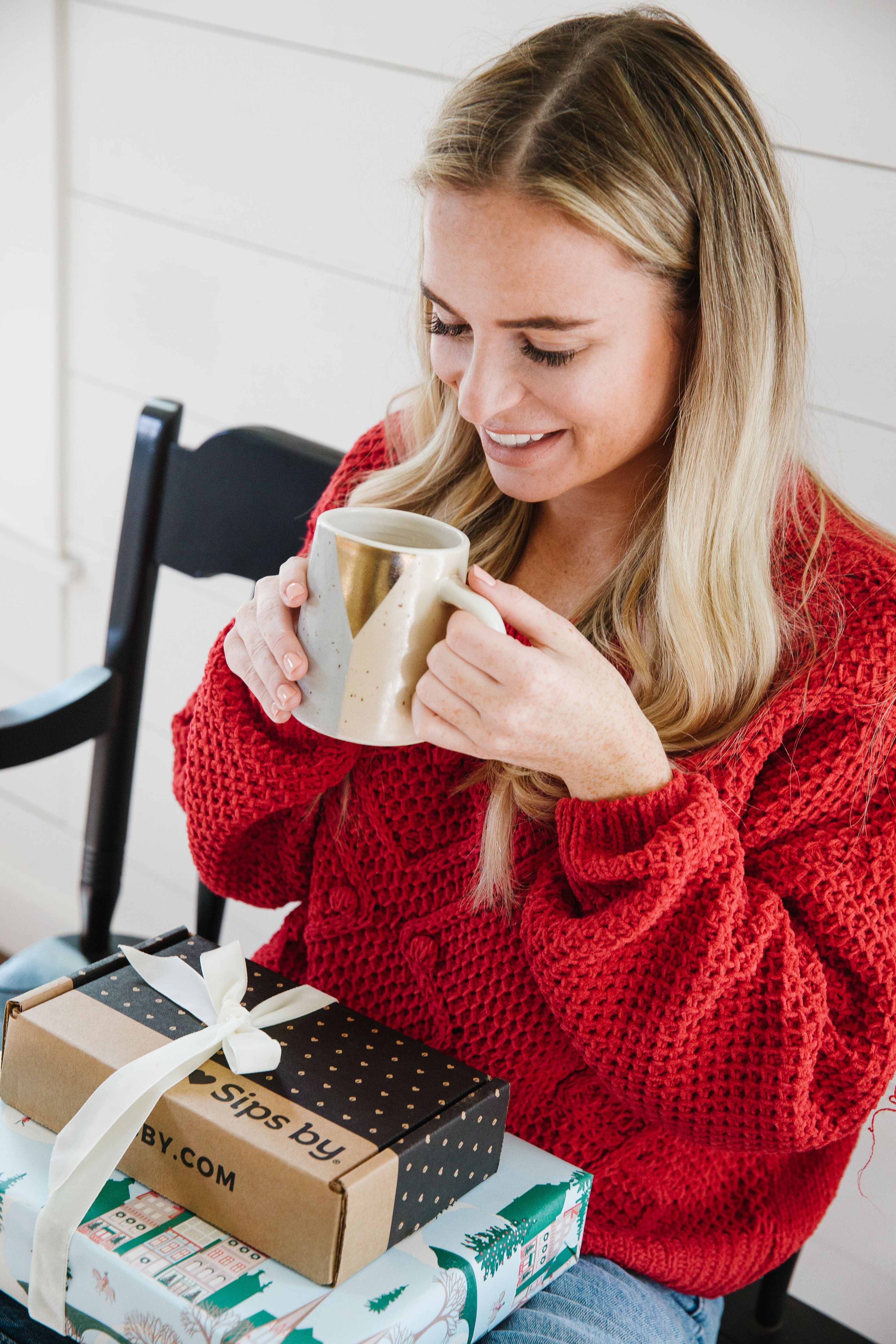 A woman in a red sweater smiling and holding a cup of tea with a stack of Sips by tea gifts in her lap
