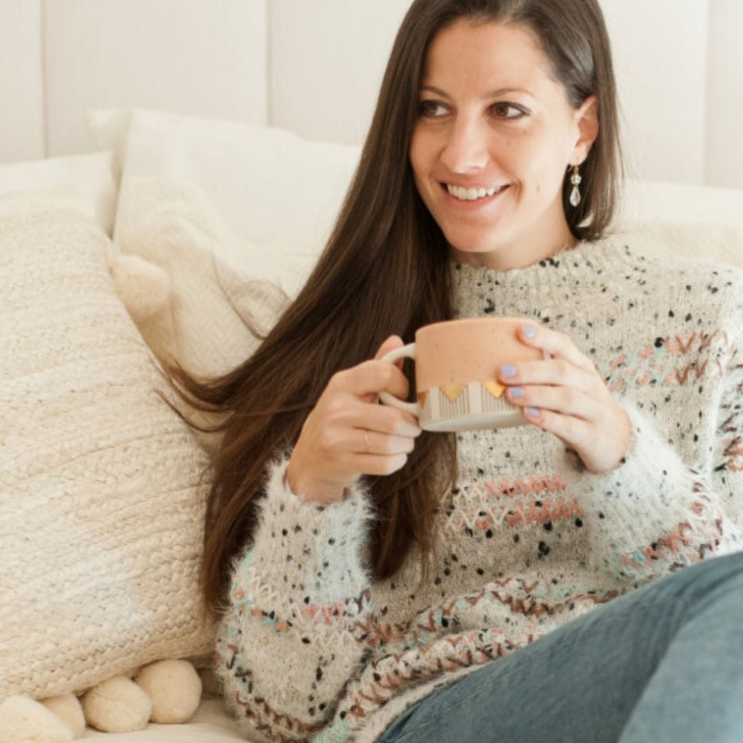 person sitting on a brown knit blanket holding a speckled mug with a chai latte
