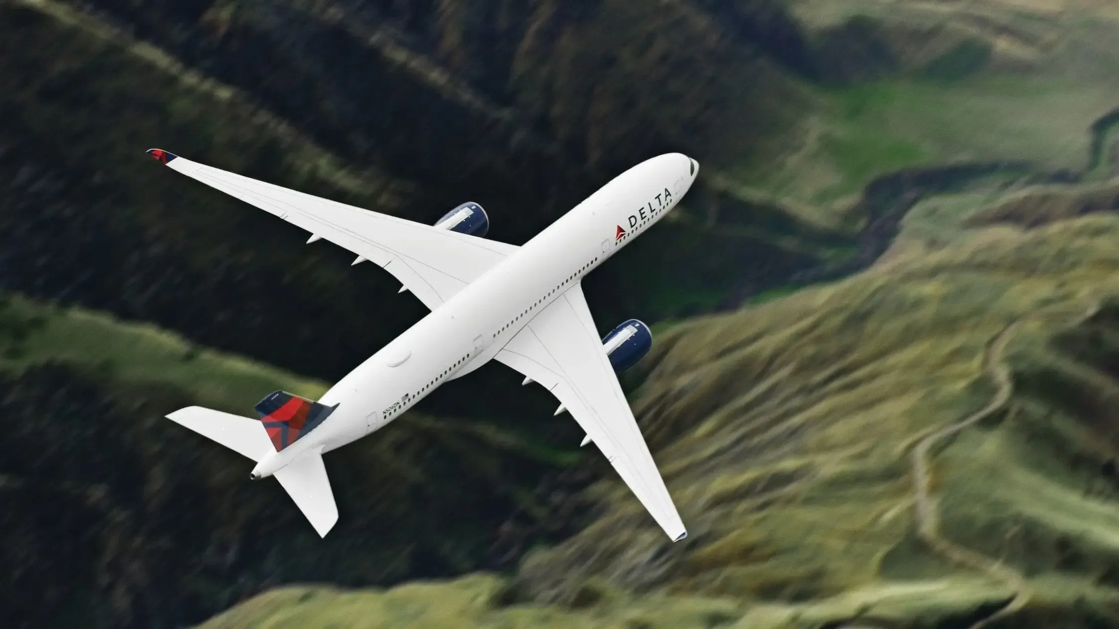 White Delta Airlines jet in flight, viewed from above, soaring over green, mountainous terrain.