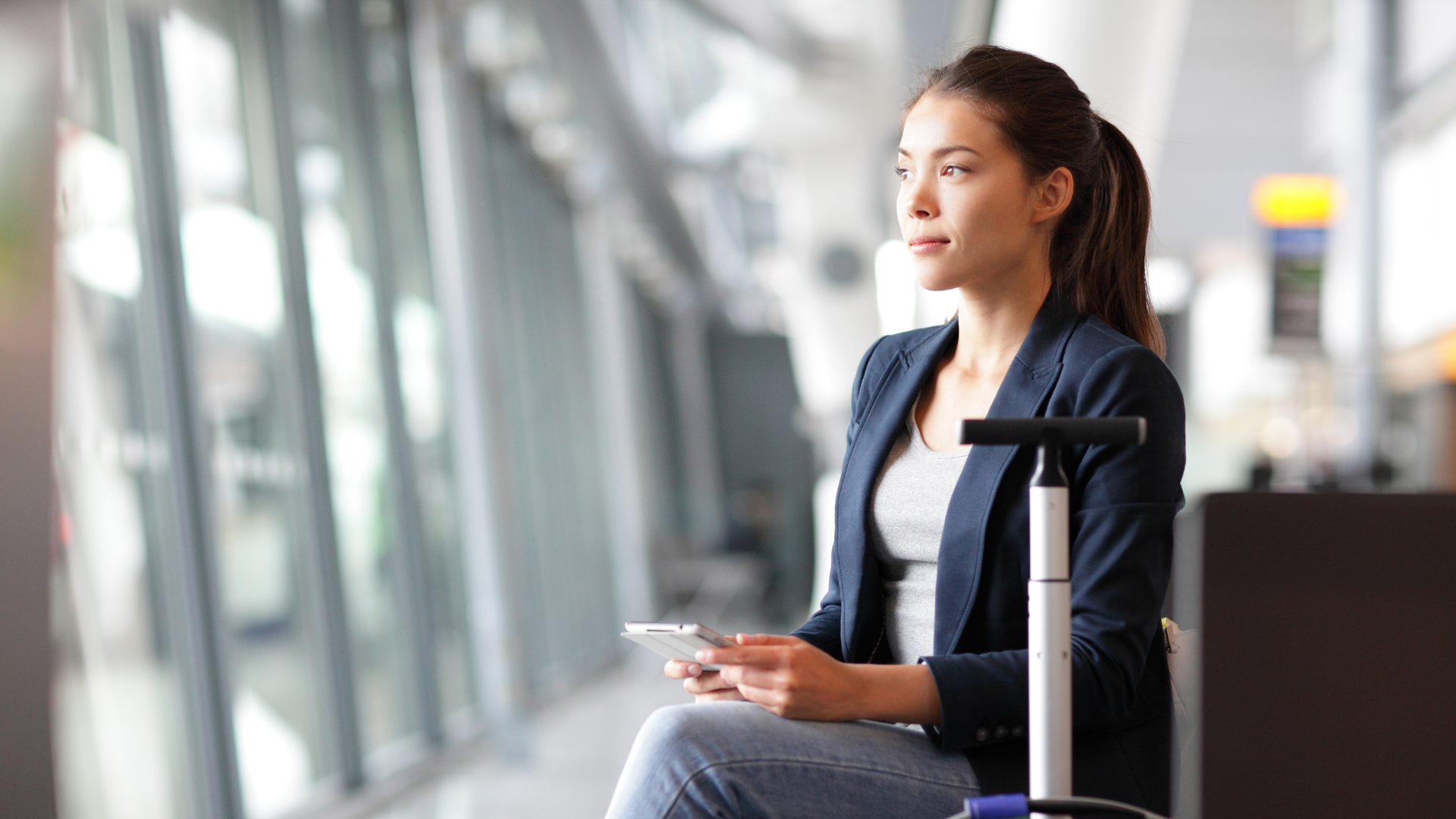 Traveler sitting in an airport terminal holding a phone, with a suitcase beside her.