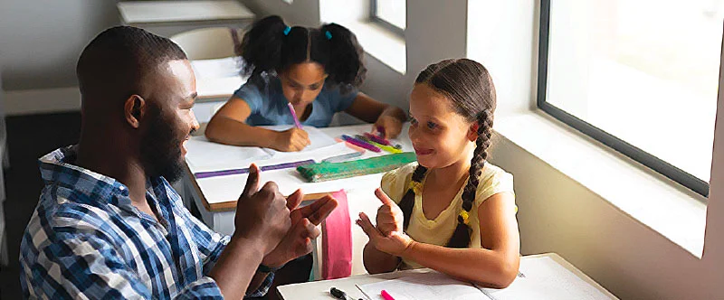 image: a black male teacher with beard showing the sign for HELP to a young brown girl with long braided pigtails who is copying him. There is another young brown girl with thick pigtails in the back writing on a paper.