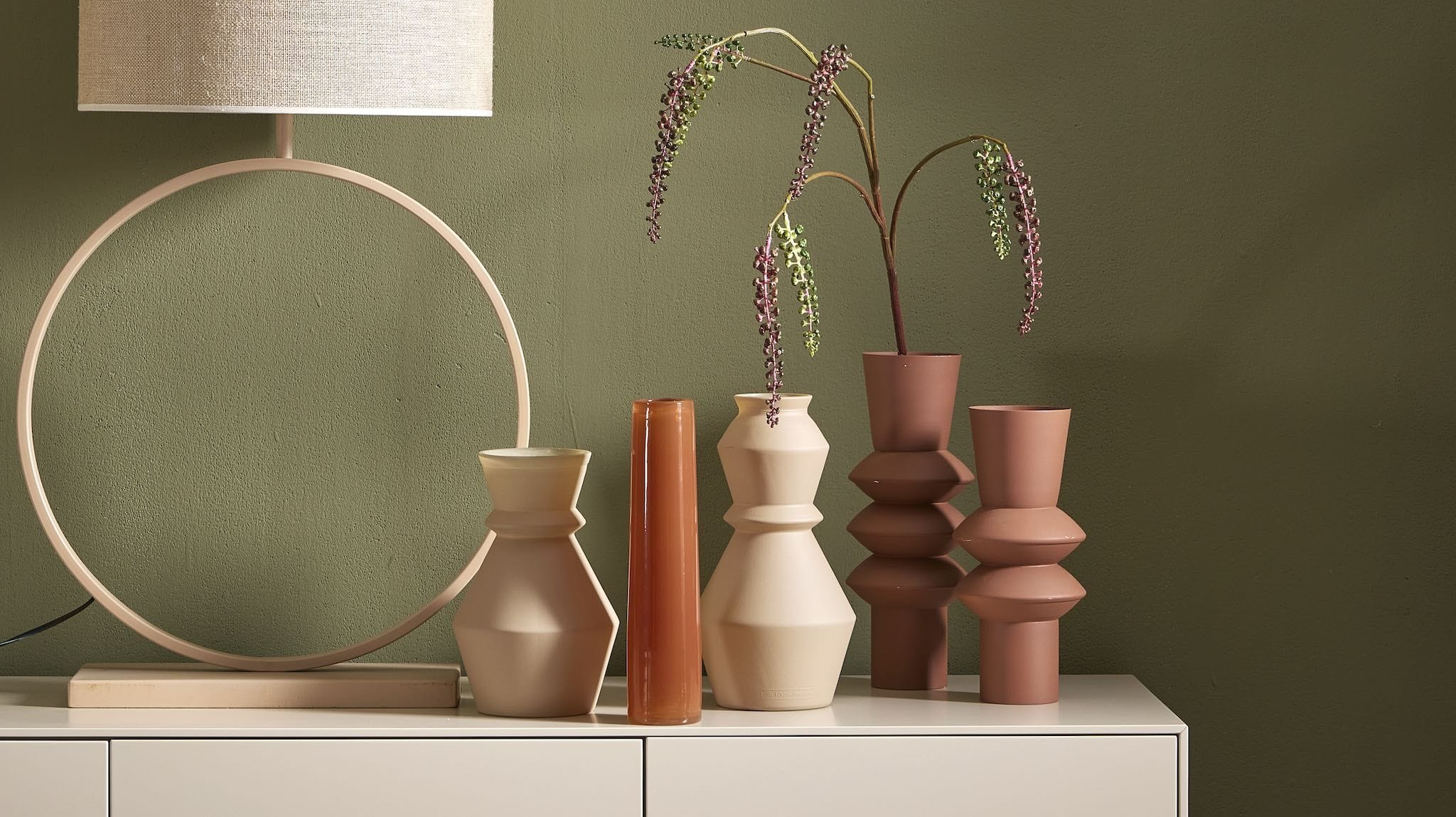 Moderne Kommode mit Dekoration, darunter eine grüne Zimmerpflanze im weißen Topf, ein Stapel Bücher, eine gelbe Designer-Vase und ein Kerzenständer vor einer olivgrünen Wand.