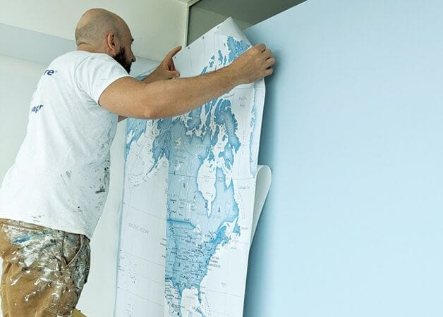 man installing a blue world map peel and stick wallpaper mural