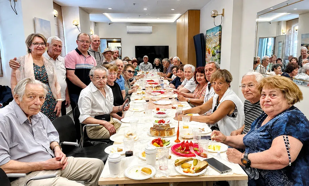 Group of older adults seated around a long table at a social gathering indoors.