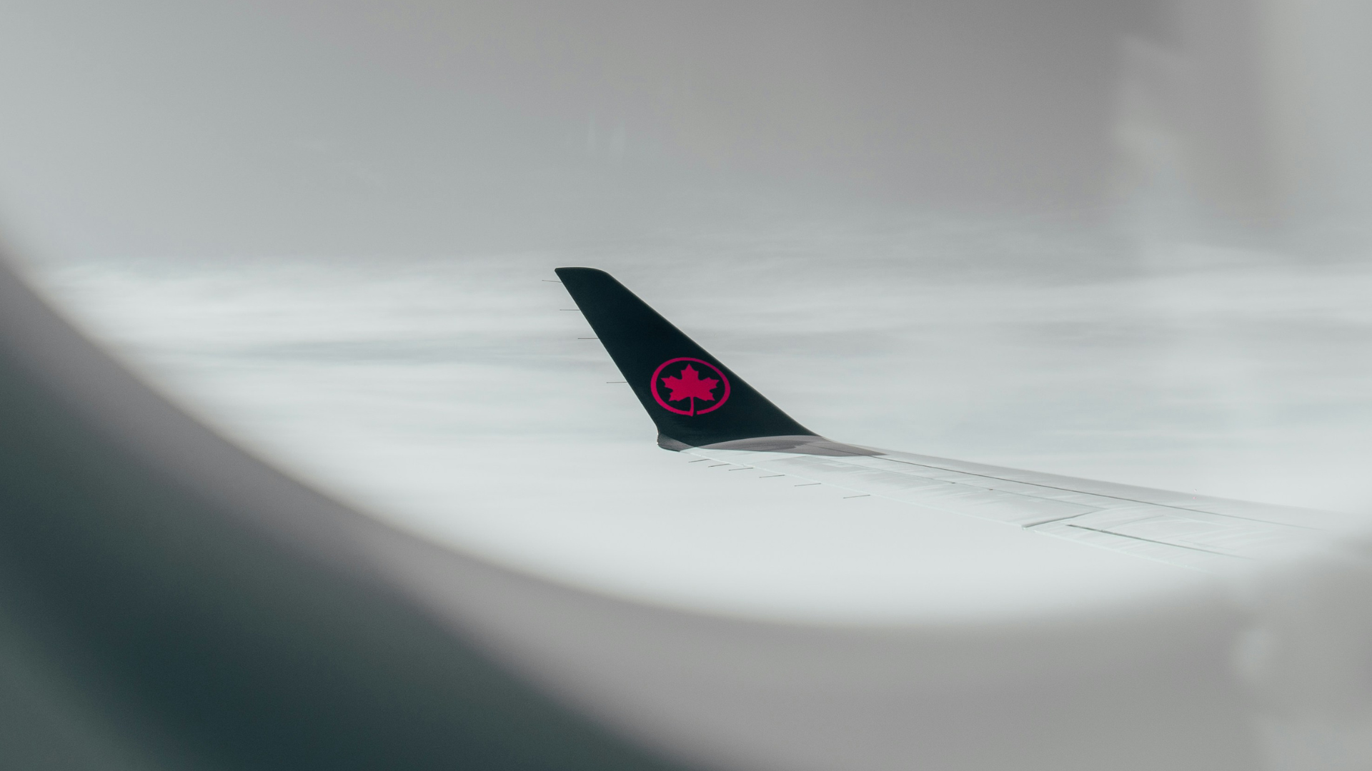 View of an airplane wing and tail with a red maple leaf logo, seen through the window against a cloudy sky.