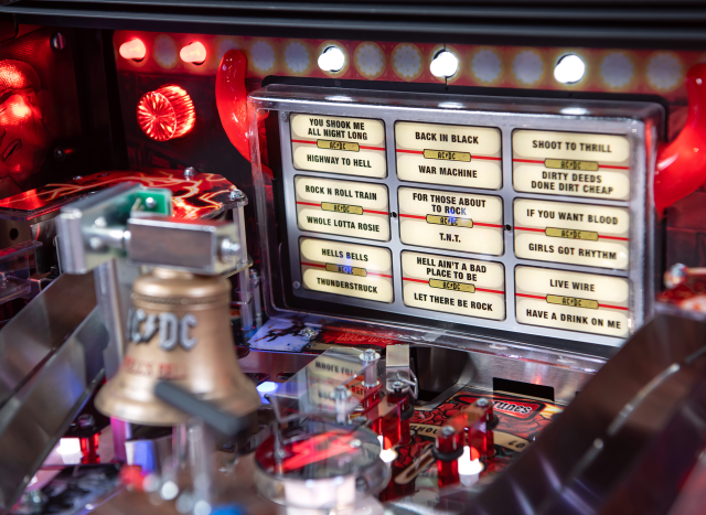 A custom sculpted bell with a illuminated song board in the background onA lower playfield with illuminated inserts on the AC/DC Remastered pinball machine by Stern Pinball.