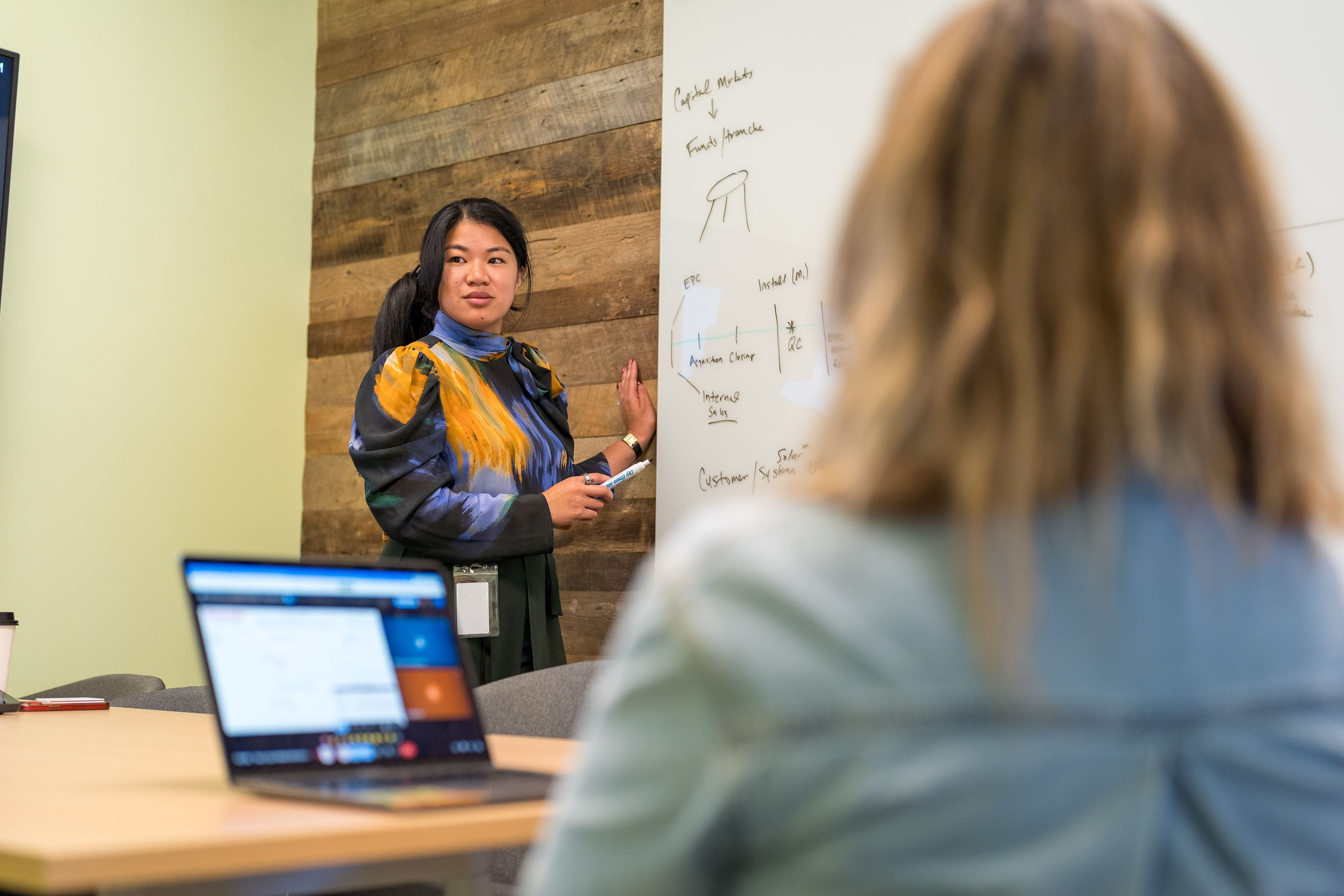 Woman looking at her laptop while sitting at a table with another woman on a whiteboard doing work.