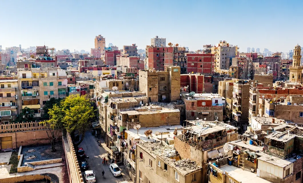 High-angle view of a dense urban neighbourhood with closely packed brick and concrete apartment buildings, rooftop satellite dishes, a narrow street below, and a mosque minaret visible on the right, possibly in Egypt.