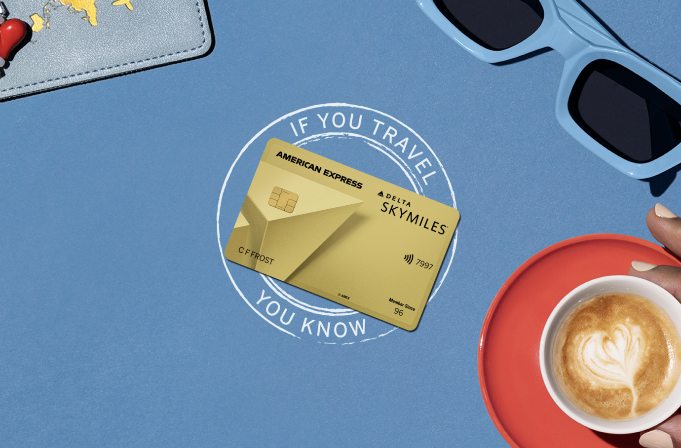 Gold-colored American Express Delta SkyMiles credit card centered on a blue background, surrounded by travel items, a coffee cup, and the text "IF YOU TRAVEL YOU KNOW."