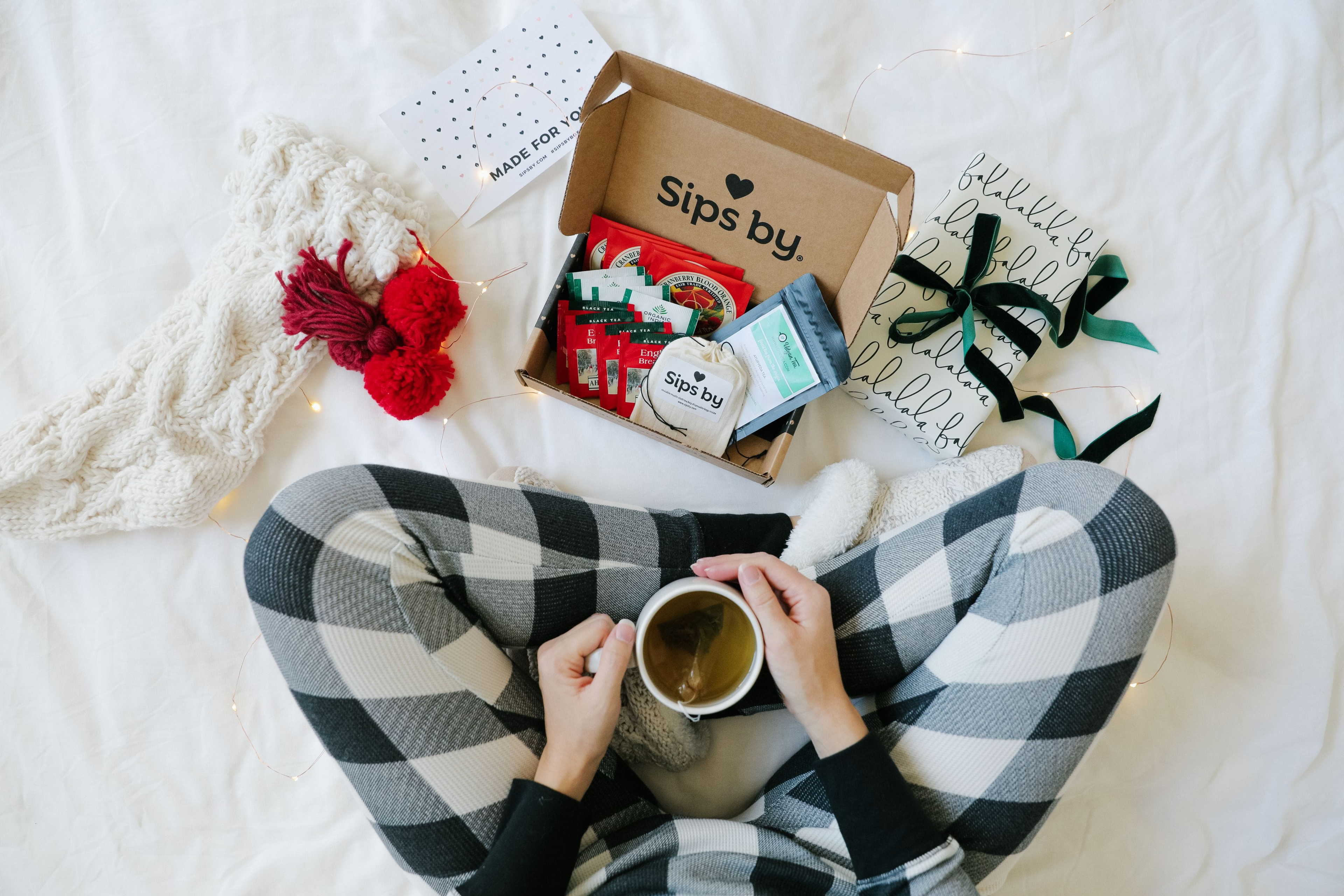 Scene of person wearing black and white flannel pajama pants, holding a mug of tea, with an open Sips by box and stocking and gifts on the floor